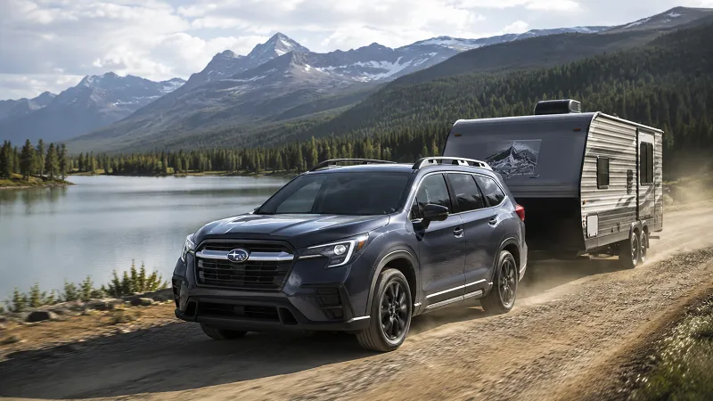 Subaru Ascent pulling a camper along a gravel road near a scenic mountain lake.